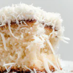 Um bolo branco coberto com glace e coco ralado, com uma fatia cortada mostrando o interior úmido do bolo.