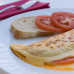 Omelete de tomate com mussarela servido em um prato branco com uma fatia de pão ao lado.