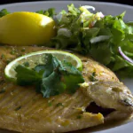 Uma tilápia assada dourada, servida com limão e salada ao lado.